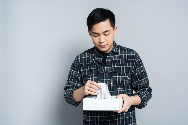 молодой азиат держит в руках пачку бумажных салфеток, страдая от насморка на сером фоне
 - Фото, изображение