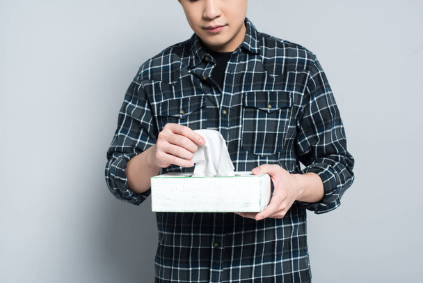 vista recortada de un joven sosteniendo un paquete de servilletas de papel mientras sufre de secreción nasal sobre fondo gris
 - Foto, Imagen