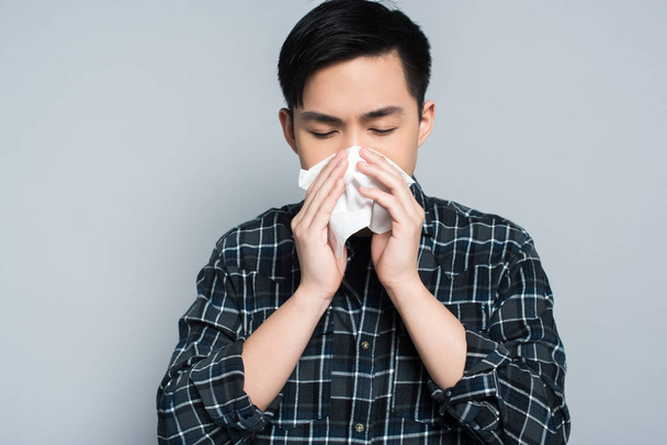 young asian man with closed eyes wiping nose with paper napkin while suffering from runny nose isolated on grey - Photo, Image