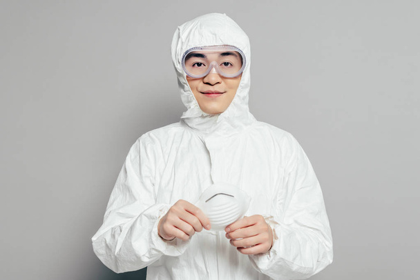 asian epidemiologist in hazmat suit holding respirator mask while looking at camera on grey background - Fotó, kép