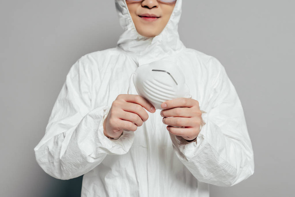 cropped view of epidemiologist in hazmat suit holding respirator mask on grey background - Fotoğraf, Görsel
