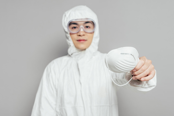 asijský epidemiolog v hazmat oblek ukazující respirátor masku při pohledu na kameru na šedém pozadí - Fotografie, Obrázek