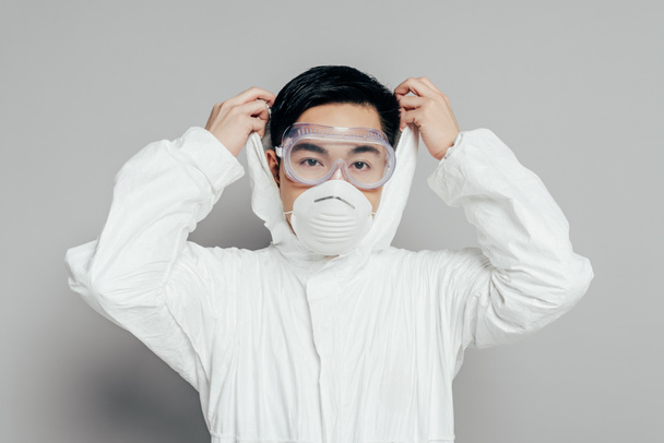 asiático epidemiólogo en hazmat traje y respirador máscara poner capucha en mientras mirando cámara en gris fondo
 - Foto, Imagen