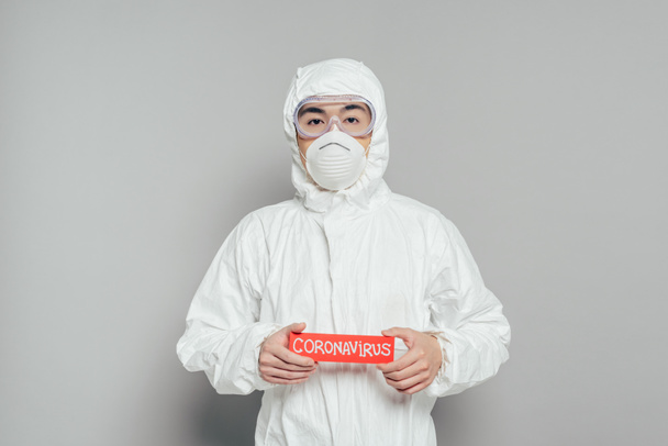 asian epidemiologist in hazmat suit and respirator mask holding warning card with coronavirus inscription on grey background - Fotoğraf, Görsel