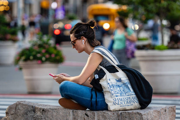 New York, USA - June 21, 2019: at the edge of the street, a woman sits on a stone and listens to music from a smartphone, blurry cityscape in the background - Foto, Bild