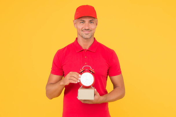 Postman delivery worker. Handsome man red cap hold alarm clock yellow background. Delivering purchase. Open hours. Time is value. Delivered to your destination. Service delivery. Courier and delivery - Foto, immagini