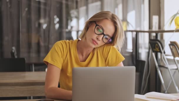 A focused blonde woman wearing eyeglasses and yellow t-shirt is working on her laptop while tilting her head in a conference hall - Záběry, video