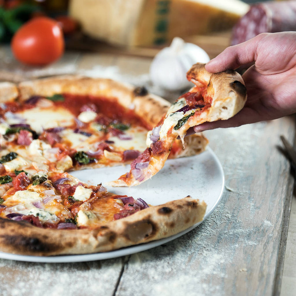 Persona recogiendo pedazo de pizza recién horneada en la mesa de madera con ingredientes
 - Foto, Imagen