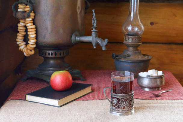 На столе стакан черного чая от старого самовара и лампа
.  - Фото, изображение