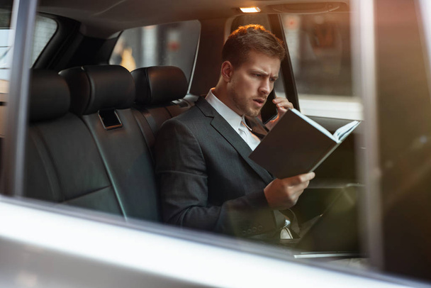 young businessman having phone conversation holds his planner while sitting in his car on the way to meetting with partners, looks busy, multitasking concept - Fotó, kép