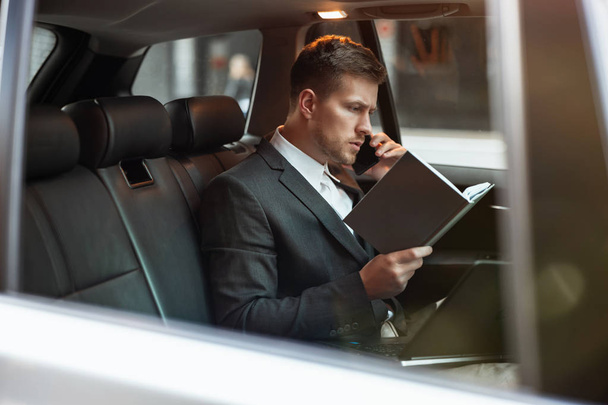 joven empresario tener conversación telefónica sostiene su planificador mientras está sentado en su coche en el camino a la reunión con los socios, concepto multitarea
 - Foto, Imagen