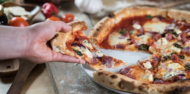 Person picking piece of fresh baked pizza on wooden table with ingredients - Photo, Image