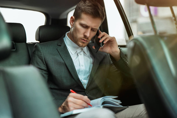 successful businessman takes notes to planner while having business phone conversation riding on back seat of car on way to meetting with partners, multitasking concept - Foto, Imagen