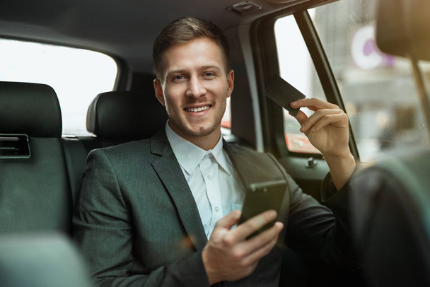 successful businessman making online payment using bank card while riding on back seat of car on way to meetting with partners, looks happy, multitasking concept - Foto, afbeelding