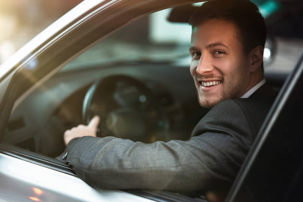 young smiling successful businessman looks happy while driving his car with open window, multitasking, big city life - Foto, afbeelding