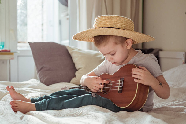 Little toddler boy in hat playing ukulele guitar at home, rustic style. Lifestyle concept - 写真・画像