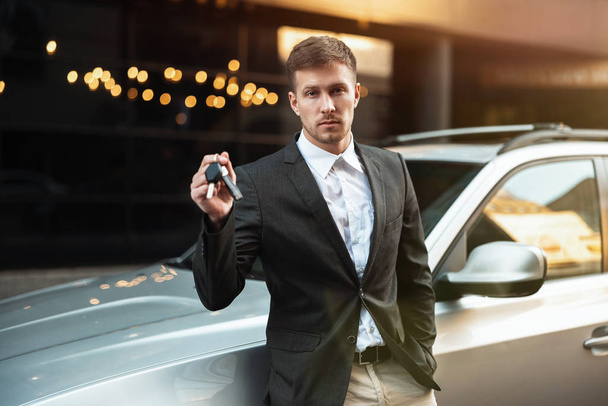 handsome businessman leaning on his parked car with keys in his hand, young and successful - Photo, Image