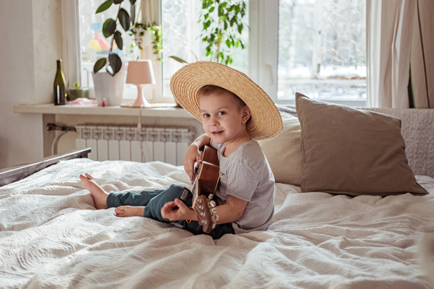Little toddler boy in hat playing ukulele guitar at home, rustic style. Lifestyle concept - Foto, afbeelding