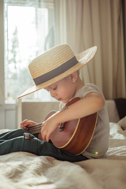 Little toddler boy in hat playing ukulele guitar at home, rustic style. Lifestyle concept - Foto, immagini