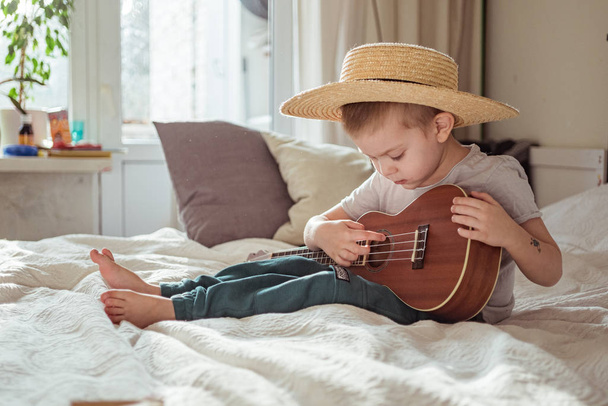 Pequeño niño en sombrero tocando la guitarra ukelele en casa, estilo rústico. Concepto de estilo de vida
 - Foto, imagen