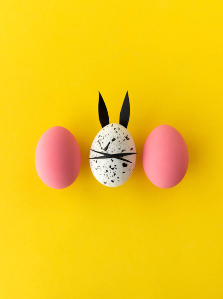 Великодня композиція з кольоровими яйцями та яйцем з вухами кролика та обличчям на жовтому фоні
 - Фото, зображення