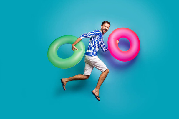 Full length photo of funky crazy guy tourist jump high running swimming hold two colorful lifebuoys circles wear striped sailor shirt shorts flip flops isolated blue color background - 写真・画像