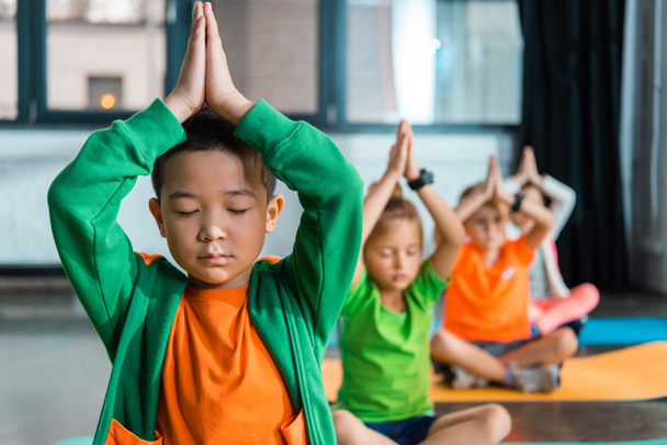 Selektywne skupienie wielokulturowych dzieci medytujących z zaciśniętymi rękami nad głowami na siłowni  - Zdjęcie, obraz
