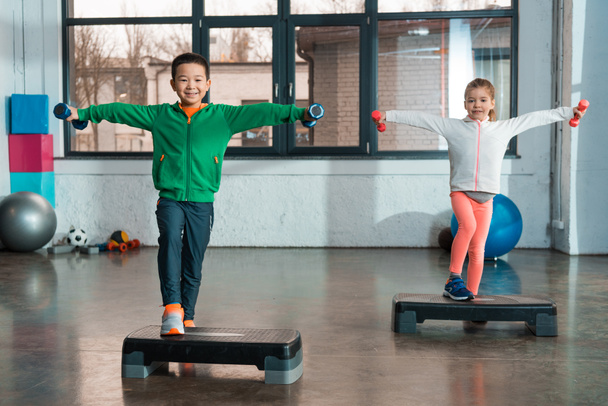 Niños multiculturales con las manos extendidas sosteniendo pesas y haciendo ejercicio en plataformas escalonadas en el gimnasio
 - Foto, Imagen