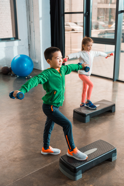 Vue en angle élevé d'enfants multiethniques avec les mains tendues tenant des haltères et faisant de l'aérobic étape dans la salle de gym
 - Photo, image