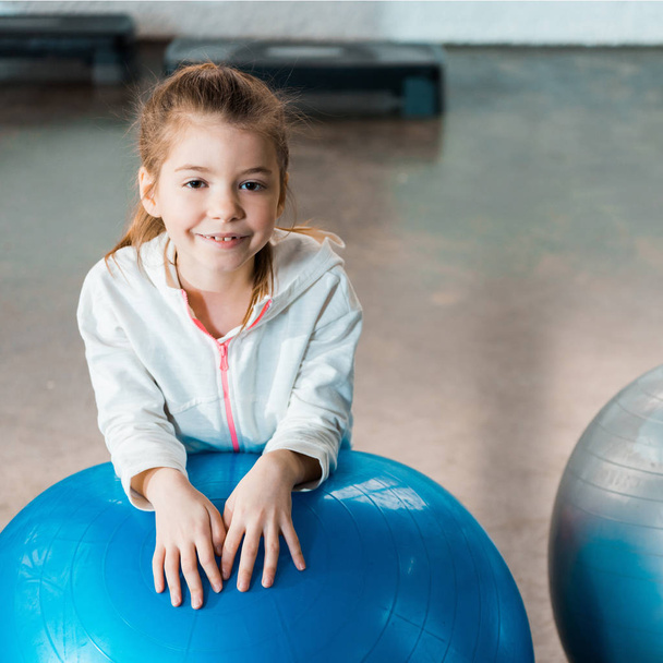 Enfoque selectivo del niño sonriendo y apoyándose en la pelota de fitness en el gimnasio
 - Foto, Imagen