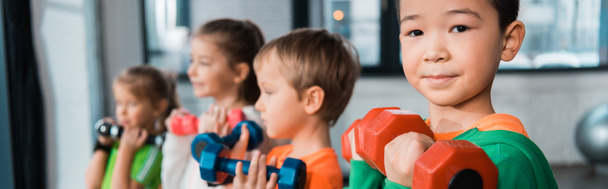 Selective focus of multiethnic children lined up, holding dumbbells in gym, panoramic shot - 写真・画像