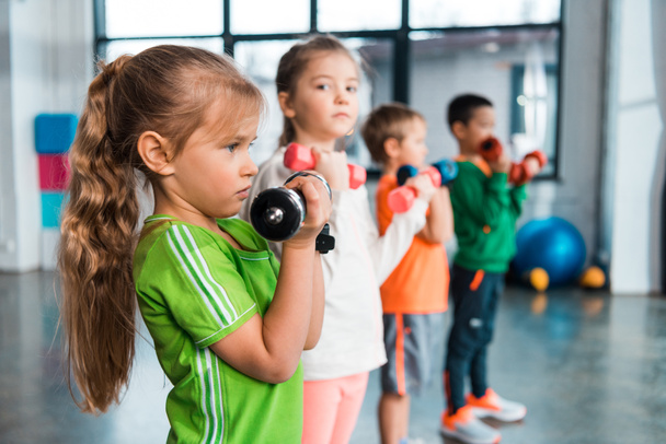 Selective focus of multicultural children lined up, holding dumbbells in sports center - Zdjęcie, obraz