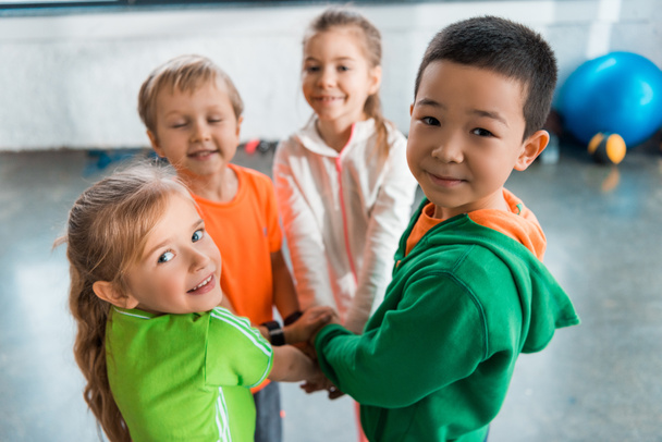 Селективный фокус мультикультурных детей, стоящих вместе в кругу с объединенными руками в спортзале
 - Фото, изображение