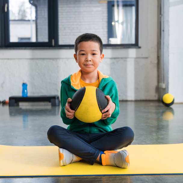Selektywne skupienie azjatyckiego chłopca trzymającego piłkę i siedzącego ze skrzyżowanymi nogami na macie fitness  - Zdjęcie, obraz