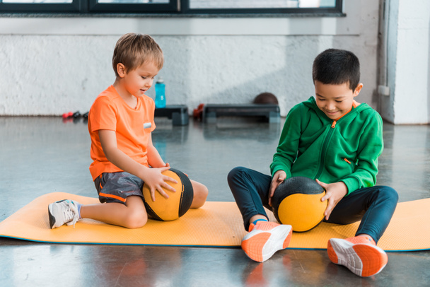 Selective focus of multicultural kids looking at balls in hands and sitting on fitness mats in gym - Fotoğraf, Görsel