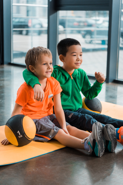 Niños multiétnicos sonriendo, abrazando y sentados con bolas en la colchoneta de fitness en el gimnasio
 - Foto, Imagen