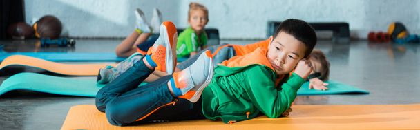 Selective focus of multicultural children lying on fitness mats in sports center, panoramic shot - Photo, Image