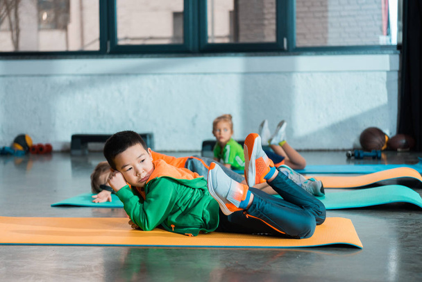 Selective focus of multicultural children resting on fitness mats in gym - Фото, изображение