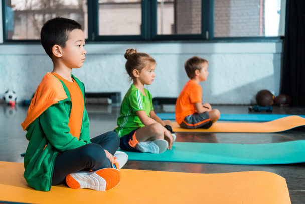 Selective focus of multicultural children with crossed legs sitting on fitness mats in sports center - Photo, Image