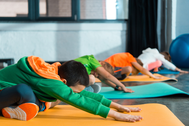 Selective focus of multiethnic children with crossed legs stretching on fitness mats in sports center - Foto, Bild