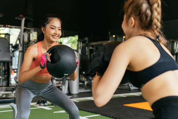 Happy young female athletic people performing squat exercises with friend and holding a medicine ball at fitness gym. Group of two confident women with healthy lifestyle working out together. - Foto, Imagem
