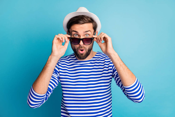 Photo of funky excited handsome guy tourist taking off cool black sun specs open mouth read discounts banner wear striped sailor shirt cap isolated blue color background - Foto, Imagen