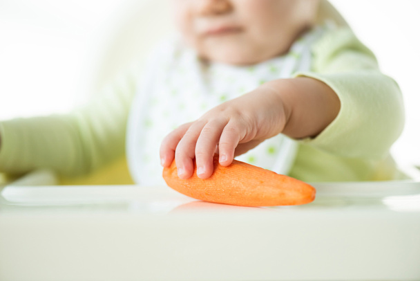 Selektiver Fokus des Babys, das Karotte hält, während es auf der Fütterung von Saibling sitzt, isoliert auf Weiß - Foto, Bild