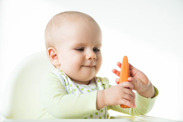 Liebenswertes Baby, das Karotte hält, während es auf einem Fütterungsstuhl sitzt, isoliert auf weißem Grund - Foto, Bild