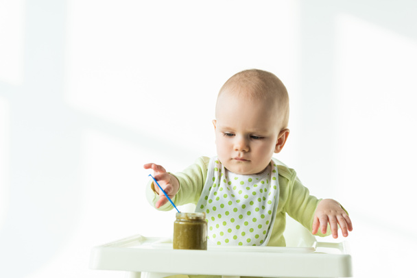 Infant holding spoon in jar of vegetable baby food on table of feeding chair on white background - Photo, Image