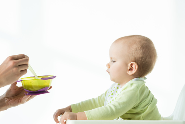 Side view of mother holding bowl of baby nutrition and spoon near infant on feeding chair on white background - Photo, Image