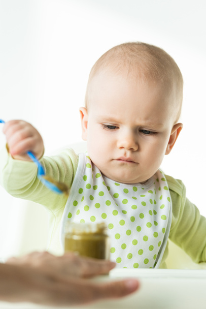 Selective focus of baby holding spoon near jar with vegetable puree and mother on white background - Photo, Image