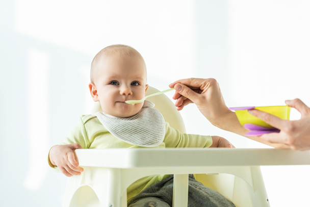 Selective focus of mother with puree feeding baby on feeding chair on white background - Фото, зображення