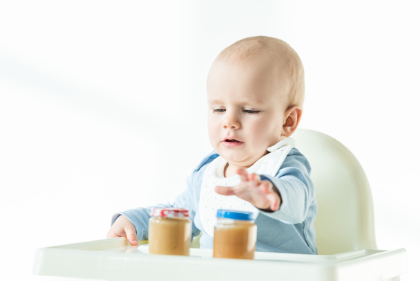 Enfoque selectivo del bebé sentado en la silla de alimentación con frascos en puré de frutas en la mesa sobre fondo blanco
 - Foto, Imagen