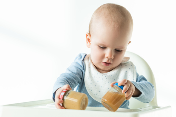 Baby holding jars of fruit puree while sitting on feeding chair on white background - Fotoğraf, Görsel